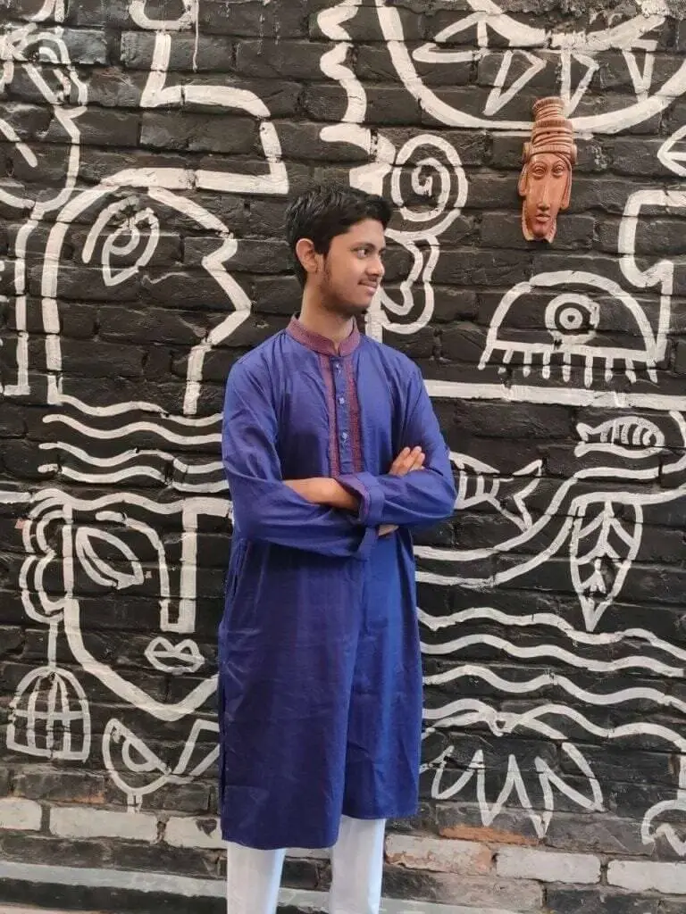 Person wearing a traditional blue kurta with folded arms, standing in front of a black brick wall adorned with white abstract artwork and a terracotta mask.