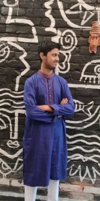 photo_2024-10-08_23-51-38-768×1024-1 (1) Person wearing a traditional blue kurta with folded arms, standing in front of a black brick wall adorned with white abstract artwork and a terracotta mask.